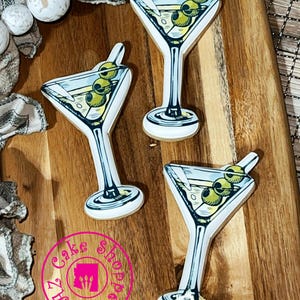 May include: Three martini-shaped cookies with olive garnishes are arranged on a wooden cutting board. The cookies have a white outline and are filled with a light yellow liquid and three green olives. The cookies are decorated with a black outline and sit on a wooden cutting board.