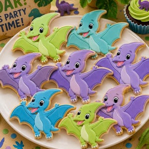 Peut inclure: Une collection de biscuits en forme de ptérodactyle décorés avec du glaçage coloré dans des tons vert lime, bleu sarcelle et violet, disposés sur une assiette blanche. Chaque biscuit présente un visage de dinosaure souriant avec des détails.