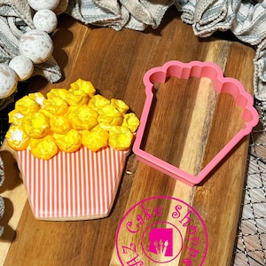 May include: A pink plastic cookie cutter shaped like a popcorn bucket. A decorated cookie with yellow popcorn and a pink and white striped bucket is next to the cutter. The cookie is decorated with a pink and white striped pattern. The cookie cutter and cookie are on a wooden surface. The image also includes a pink circle with the text "Cake Supply" and "Est. 2011".