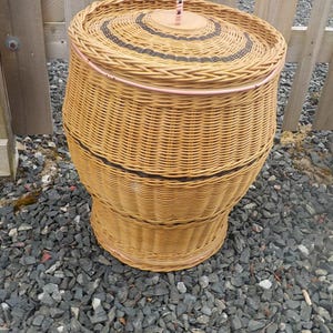 May include: A light brown wicker laundry basket with a lid. The basket has black and pink trim. The lid has a wooden handle. The basket is on a bed of gray gravel.