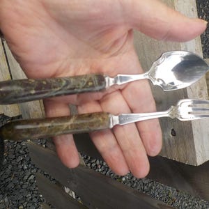 May include: A silver spoon and fork set with decorative edges and handles made of a speckled, dark brown and green material. The spoon has a rounded bowl, and the fork has three tines. The set is displayed on a wooden surface.