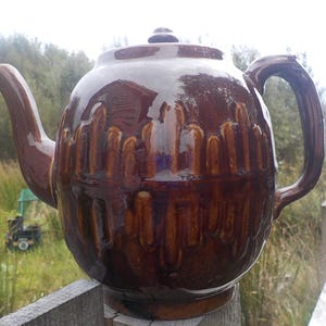 May include: A brown ceramic teapot with a glossy finish. The teapot has a rounded body, a curved spout, and a loop handle. The surface is decorated with vertical, textured patterns. The lid has a small knob. The teapot is sitting on a wooden surface.