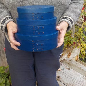 May include: Three oval, dark blue wooden boxes, stacked together. Each box has a lid and is decorated with small, evenly spaced, brass-coloured rivets. The boxes are held by a person wearing a grey sweater and navy blue trousers.