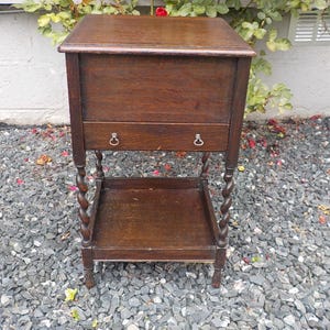 May include: A dark brown wooden side table featuring a rectangular top, a single drawer with two small metal ring pulls, and a lower open shelf. The table is supported by four legs with a twisted barley-twist design and stands on small feet.