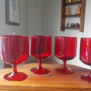 May include: Four vintage red wine glasses with a rounded bowl and a short stem. The glasses are made of a transparent red glass and are arranged on a wooden surface. The glasses are a vibrant red colour.