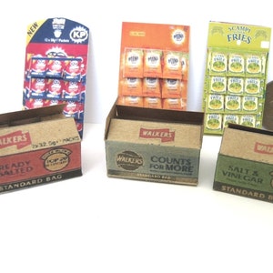 Puede incluir: Tres cajas de cartón de patatas fritas Walker's, con los sabores Ready Salted, Salt & Vinegar y Counts for More. Las cajas están sobre una superficie blanca, con una moneda y dos expositores de aperitivos en el fondo.