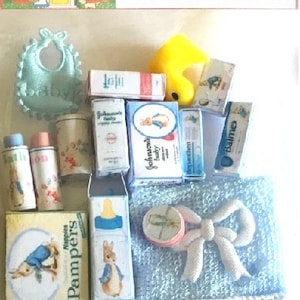 May include: A collection of miniature baby care products, such as bottles, powder, and a bib, are displayed on a light blue towel. The image includes a yellow rubber duck and packaging with the text "Johnson's baby" and "Pampers". The text "Hand crafted Dolls house Miniatures" is at the top.