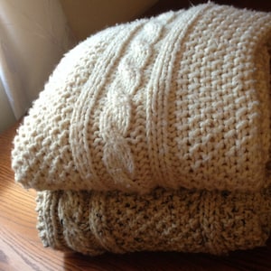 May include: Two folded, chunky knit blankets in cream and tan. The top blanket features a cable knit design, while the bottom blanket has a textured pattern. The blankets are stacked on a wooden surface, suggesting a cozy, home decor theme.
