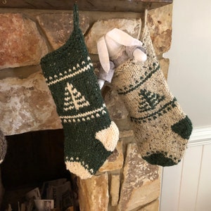 May include: Two hand-knitted Christmas stockings featuring a tree motif. One stocking is dark green with cream accents, and the other is cream with dark green accents. A small fabric rabbit toy with long ears is tucked inside the cream stocking.