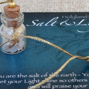 May include: A gold necklace featuring a small glass bottle filled with salt and gold flakes. The bottle is attached to a brown wooden bead with a gold cord. The necklace is displayed on a blue background with the text "Holyland's Salt & Light" and a bible verse.