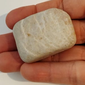Może przedstawiać: Gładki, podłużny, jasno zabarwiony kamień z subtelnymi żyłkami i zaokrąglonym kształtem. Kamień ma naturalny, zwietrzały wygląd, z różnicami w kolorze i fakturze. Kamień jest trzymany w otwartej dłoni.