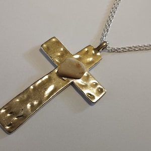 Pode incluir: Um pendente de cruz dourada com textura martelada, apresentando uma pequena pedra clara no centro. A cruz pende de uma delicada corrente prateada, criando um colar simples, mas elegante.
