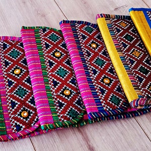 以下が含まれることがあります： 幾何学模様と金色のアクセントが施された、カラフルな刺繍の布製クラッチが6つセットになっています。クラッチは木製の表面に一列に並んでいます。