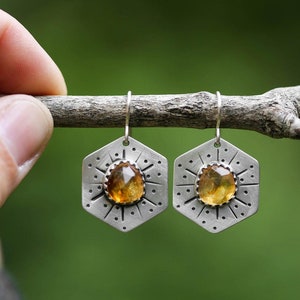 Puede incluir: Un par de pendientes de plata con un diseño hexagonal y una piedra preciosa amarilla en el centro. Los pendientes están colgados de una rama.