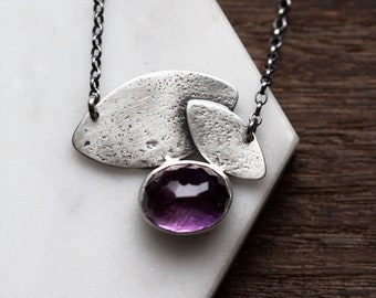 May include: A silver necklace with a textured pendant featuring three overlapping oval shapes and a round amethyst gemstone.