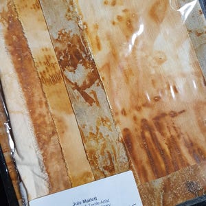 May include: A pack of fabric strips featuring rust-dyed patterns in shades of orange, brown, and cream. The packaging includes a white label with the text Jule Mallett, Mixed Media & Textile Artist, Handmade - Less Ordinary, julemallett.uk, Rust Dyed Fabric Sample Pack.