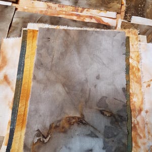 May include: A collection of rectangular fabric swatches featuring botanical eco-prints. The pieces display earthy tones including muted greys, deep browns, and golden yellows with organic leaf patterns and mottled textures on the textile surfaces.