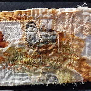 May include: A rectangular textile art piece featuring layered fabric scraps in shades of cream, beige, and brown. The composition includes hand-stitched embroidery of orange flowers and green stems, with visible frayed edges and decorative cross-stitching.