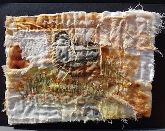 May include: A rectangular textile art piece featuring layered fabric scraps in shades of cream, beige, and brown. The composition includes hand-stitched embroidery of orange flowers and green stems, with visible frayed edges and decorative cross-stitching.