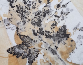 May include: A white fabric textile featuring a botanical print with dark charcoal black fern leaf impressions and soft golden-yellow watercolor-style ink splatters and stains across the surface, creating a textured and organic visual pattern.