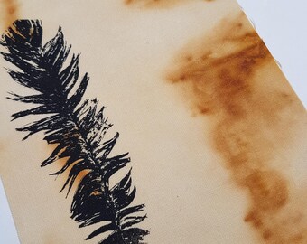 May include: A rectangular piece of beige fabric featuring a black screen-printed feather graphic on the left side. The fabric displays irregular, mottled brown stains across its surface, creating a textured, organic appearance throughout the material.