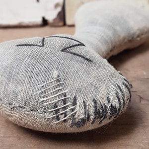May include: A handmade gray fabric doll with black stitching. The doll has a simple face with a stitched-on smile and a small, rounded body.