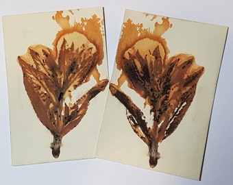 May include: Two cream-colored cards each feature a symmetrical brown ink blot resembling a stylized flower. The ink has a gradient effect, with darker brown details and lighter tan hues. The cards are angled towards each other.