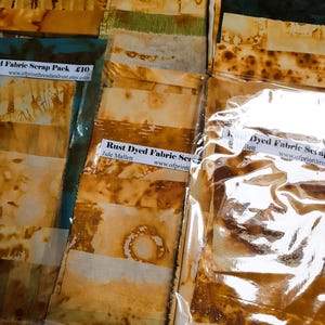 May include: Rust-dyed fabric scrap packs in shades of brown, tan, and green, packaged in clear plastic bags. The fabric pieces have a mottled appearance. Labels read "Rust Dyed Fabric Scrap Pack".