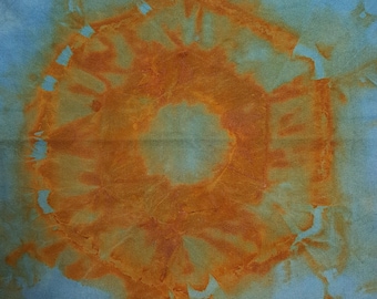 Puede incluir: Una pieza de tela cuadrada con un color base azul claro y un gran patrón circular de teñido anudado en color óxido en el centro. Los bordes de la tela están rematados con un fleco corto de color azul claro a juego en los lados superior e inferior.