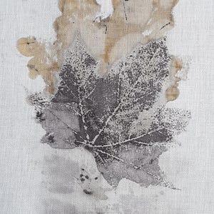 May include: A close-up of white fabric featuring a large leaf print in grey and brown tones. The leaf's intricate details are visible, with a textured effect. The design evokes a natural, botanical theme.