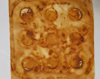 May include: A square piece of fabric featuring a grid of nine circular, coffee-stained patterns. The textile displays varying shades of brown, ranging from light tan to deep amber, with darker, concentrated rings around each circular shape.