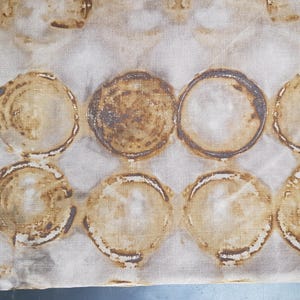 May include: A length of beige fabric featuring a repeating pattern of large, brown, circular designs. The circles have a textured look, with darker brown borders and lighter brown centres. The fabric has a raw, unfinished edge.