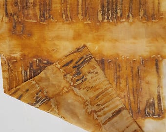 May include: A rectangular piece of fabric featuring a tie-dye pattern in shades of golden yellow, amber, and dark brown. The textile displays vertical, linear markings and organic, mottled patches, with one corner folded over to reveal the underside.