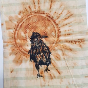 May include: A rectangular fabric piece featuring a black ink illustration of a crow standing in front of a large, circular, rust-coloured sunburst design. The background consists of light cream and pale green horizontal stripes throughout.