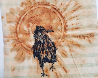 May include: A rectangular fabric piece featuring a black ink illustration of a crow standing in front of a large, circular, rust-colored sunburst design. The background consists of light cream and pale green horizontal stripes throughout.