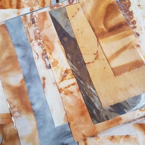 May include: A collection of rectangular fabric strips featuring a mix of rust-coloured botanical-style stains and solid slate-grey tones. The pieces are arranged in a layered, overlapping composition with visible raw edges and varied textures.