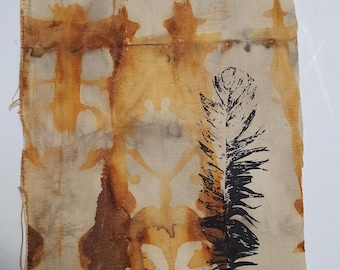 May include: A rectangular piece of fabric featuring a tan base color with abstract, symmetrical rust-colored patterns created by tie-dye or botanical printing. A single, detailed black ink print of a feather is positioned on the right side.