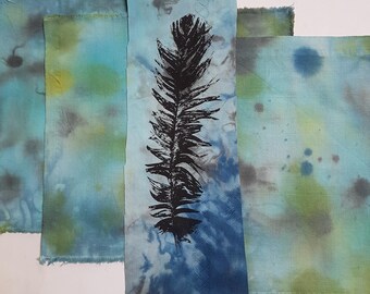 May include: A collection of rectangular fabric pieces featuring a tie-dye pattern in shades of blue, green, and grey. The central vertical strip displays a bold, black ink-stamped graphic of a single feather with detailed barbs and a central shaft.