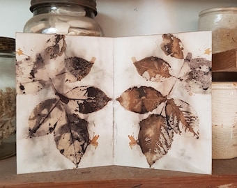 May include: A folded card featuring a botanical print of leaves in shades of brown and beige. The card is displayed on a wooden shelf, with various jars and containers in the background. The overall aesthetic is rustic and natural.