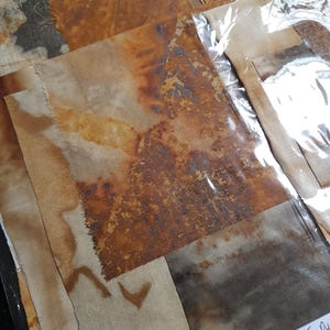 May include: A collection of rectangular fabric scraps featuring various rust and tea-dyed patterns in shades of brown, tan, and grey. The pieces are layered and partially enclosed in a clear plastic sleeve with a label reading Scrap Pack Rust + Tea Dye.