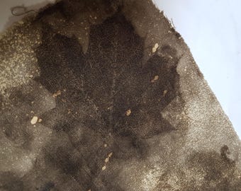 May include: A piece of fabric featuring a dark brown botanical print of a maple leaf. The textile displays a textured, mottled appearance with lighter beige speckles and patches throughout the surface, creating a natural, earthy aesthetic.