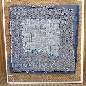 May include: A square textile art piece featuring layers of blue denim fabric with visible white stitching. The centre displays a woven denim patch with frayed edges, mounted on a rectangular board covered in a tan, textured burlap-like material.