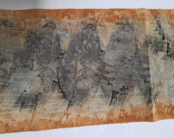 May include: A rectangular strip of fabric featuring a botanical print with three distinct dark gray leaf silhouettes centered on a textured, warm orange-brown background. The edges of the fabric show a crinkled, uneven dyed pattern throughout.
