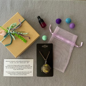 Op de afbeelding: Een bruine doos met een groen-wit lint, een klein flesje etherische olie, een kleine groene bal, een paarse organza zak en een zilveren ketting met een ronde, sierlijke chime ball hanger. De ketting ligt op een zwarte kaart met de tekst "AROMATHERAPY DIFFUSER & CHIME BALL ANGEL CALLER NECKLACE SET".