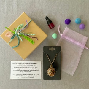 May include: An aromatherapy diffuser and chime ball necklace set. The set includes a gold-colored filigree locket pendant, a small bottle, a gift box with ribbon, a sheer pouch, and colorful felt balls.