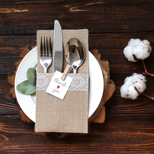 Può includere: Una mise en place rustica con un tovagliolo di juta, un bordo in pizzo bianco e un biglietto di ringraziamento. Il tovagliolo contiene una forchetta, un coltello e un cucchiaio. La mise en place è su un piatto bianco con una base in legno.