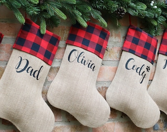 May include: Five personalized burlap Christmas stockings with red and black buffalo plaid trim. The stockings are labeled "Dad", "Olivia", "Cody", and "Mom".