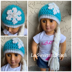 May include: A turquoise crochet hat with a white snowflake design and a silver trim. The hat is worn by a doll with a white shirt that says "ado" and a blue skirt. The doll has long white and silver braided hair.