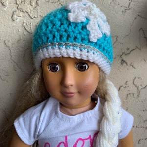May include: A blue crocheted hat with a white snowflake design and a silver trim. The hat is worn by a doll with blonde hair and a white braid. The doll is wearing a white shirt with the word "ador" written in pink.