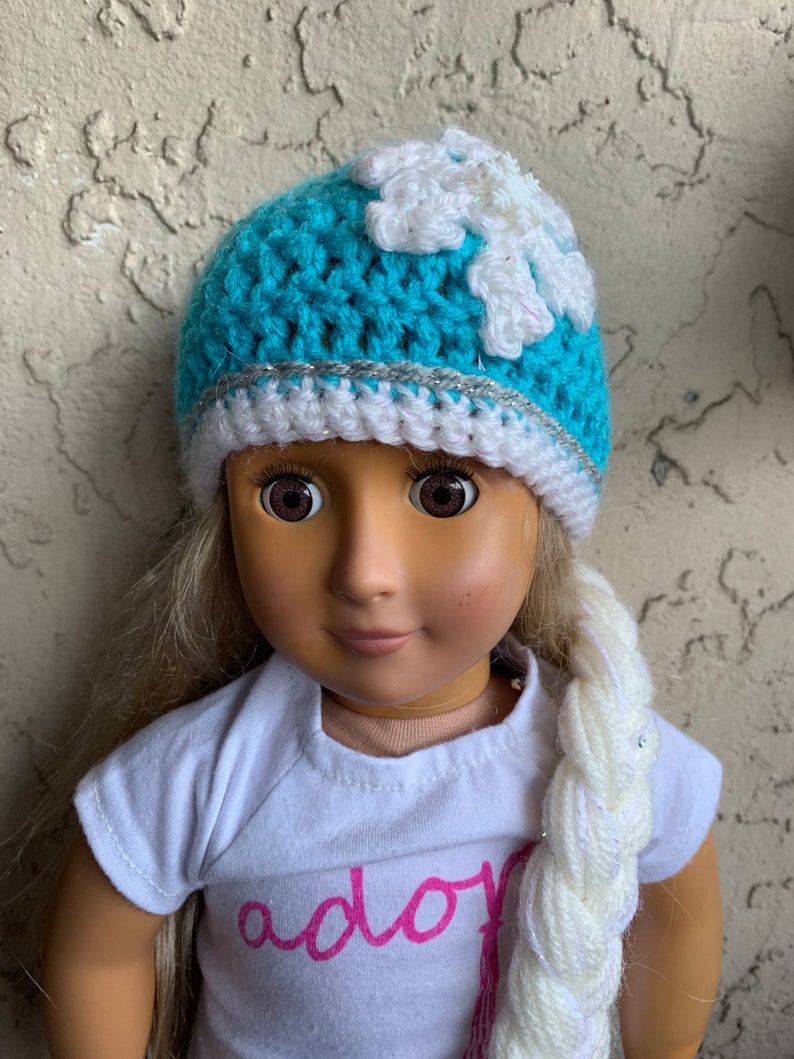 May include: A blue crocheted hat with a white snowflake design and a silver trim. The hat is worn by a doll with blonde hair and a white braid. The doll is wearing a white shirt with the word "ador" written in pink.